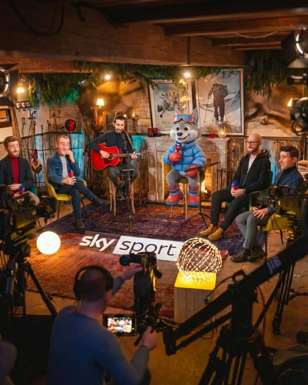 Il personaggio Asso seduto con il microfono in mano su un set televisivo Sky Sport, fra gli ospiti in studio e le telecamere di ripresa