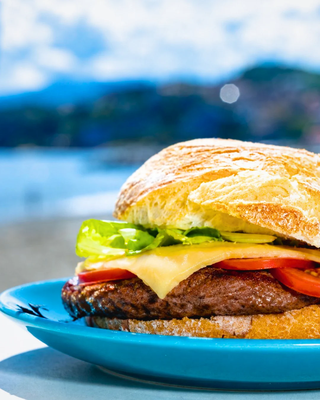 Panino con hamburger, formaggio e pomodoro su un piatto azzurro, con il mare sfocato alle spalle