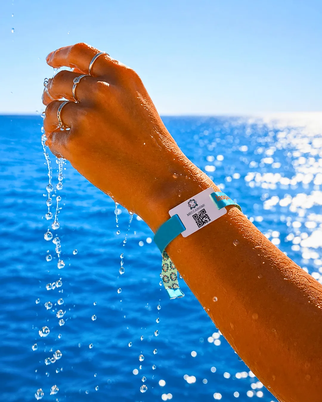 Braccio che esce dall'acqua con il braccialetto e il codice QR dell'evento al polso, mare blu alle spalle